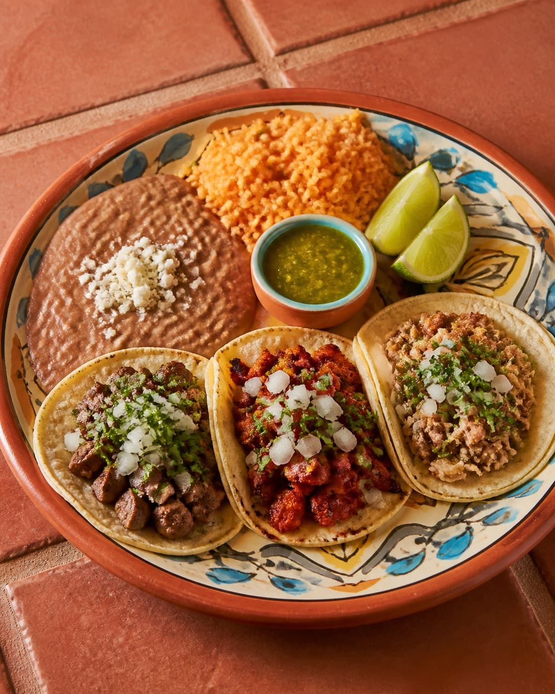 Three street tacos with rice, beans, green salsa, and lime on a painted clay plate